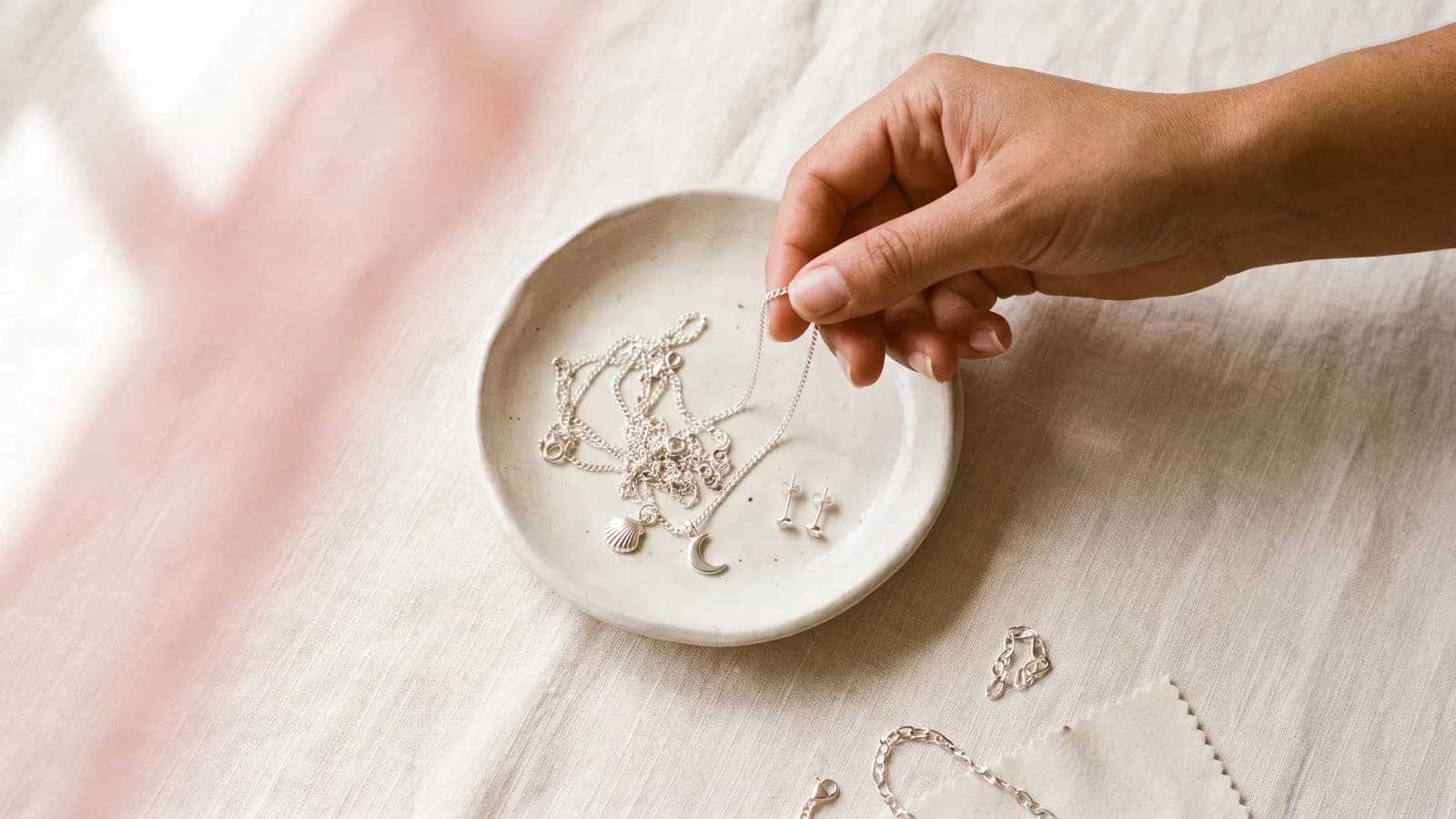 Fine sterling silver chains and charms in a shallow dish on cream linen, one chain being lifted out
